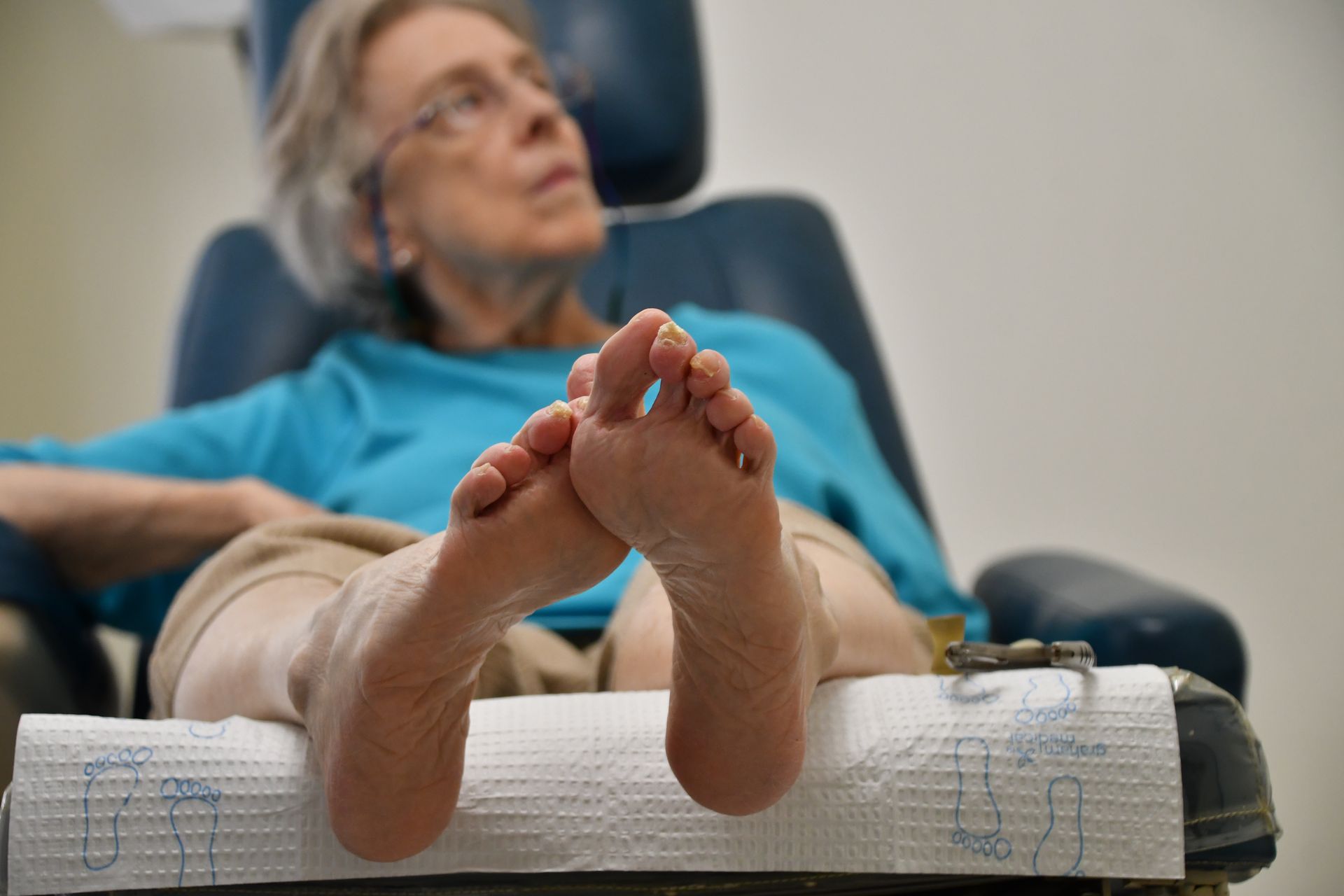 Woman in blue shirt reclines in chair with feet propped up, likely at a podiatrist's office.