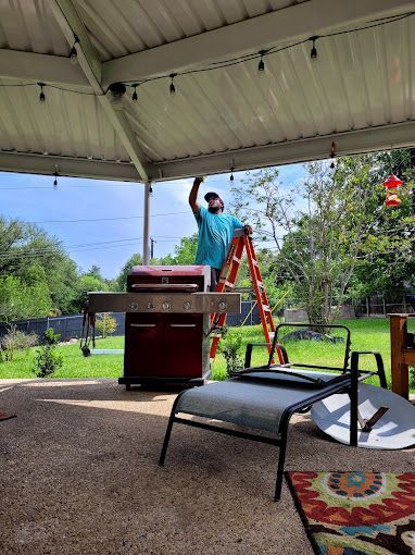 AquaWorks Rain Gutters - Man on a ladder hanging string lights under a patio roof in a backyard, near a grill.