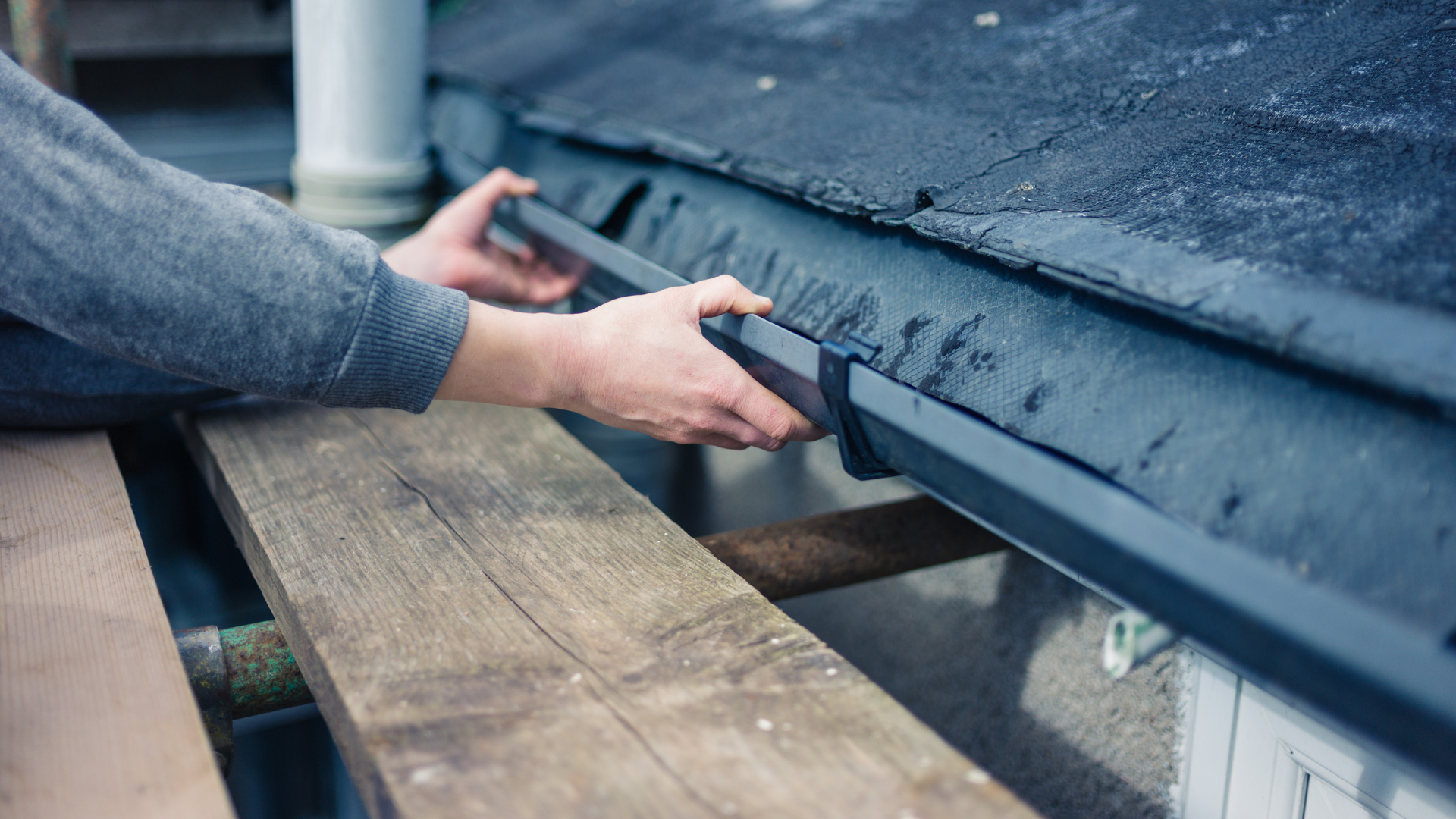 AquaWorks Rain Gutters - Person installing a black gutter on a roof, wooden plank in foreground.