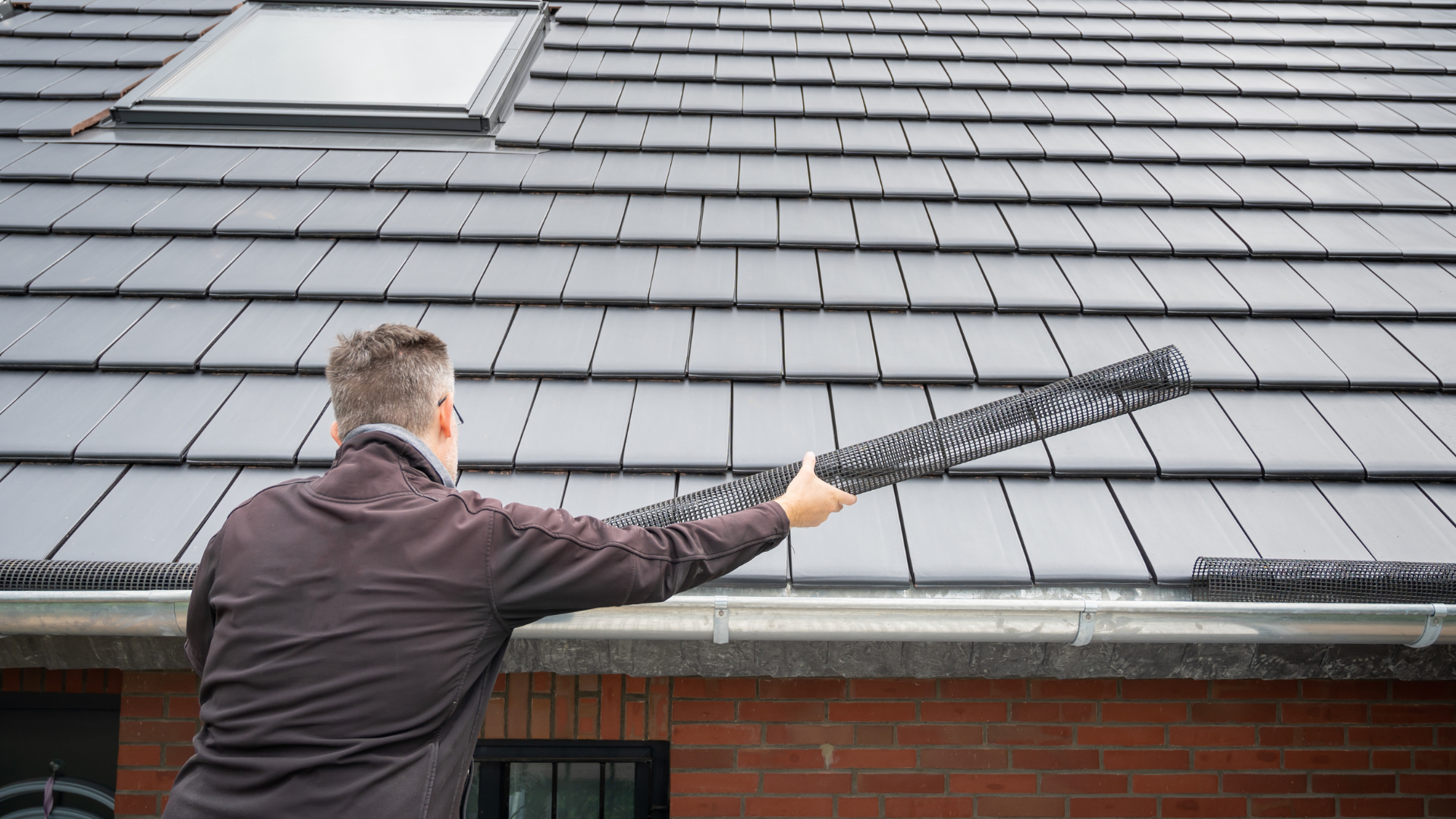 AquaWorks Rain Gutters - Person on roof installing gutter guard. Gray roof tiles, light-colored gutter, brick wall.