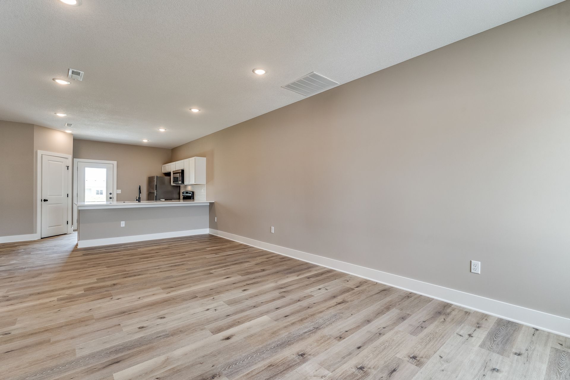 An empty living room with hardwood floors and a kitchen in the background.