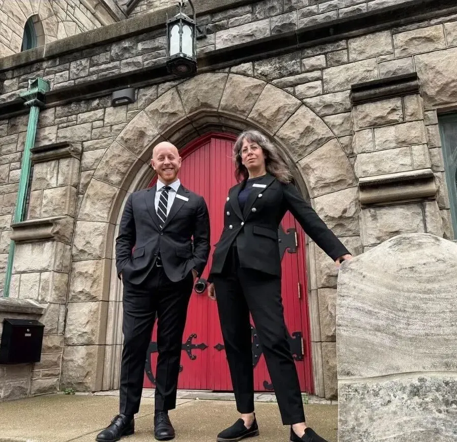 Two people in black outfits stand in front of a stone building with a red arched door.