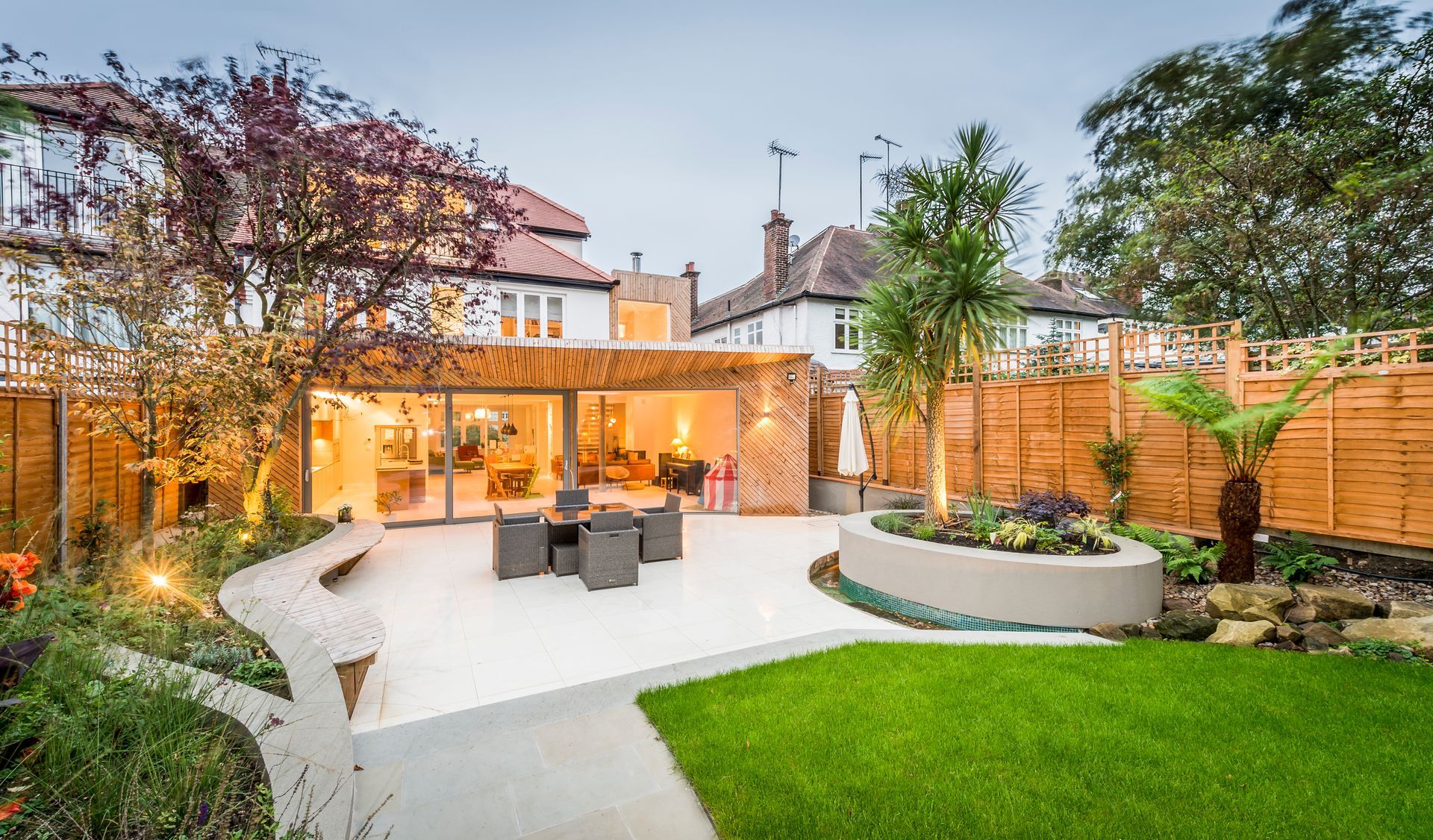 Backyard patio with modern design: concrete, grass, trees, seating, and a house with large windows.