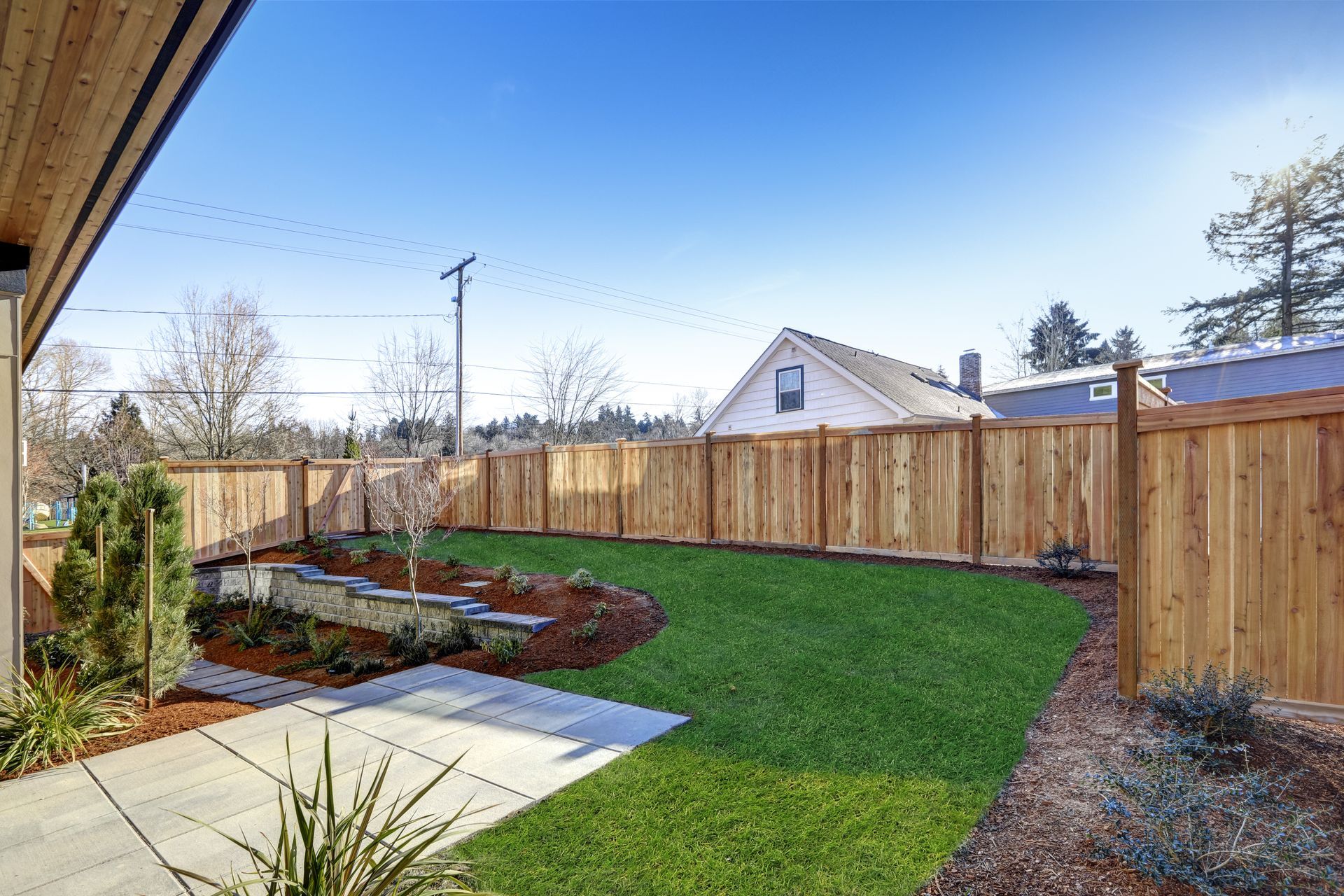 Backyard with a wooden fence, green lawn, flower beds, and a patio under a blue sky.