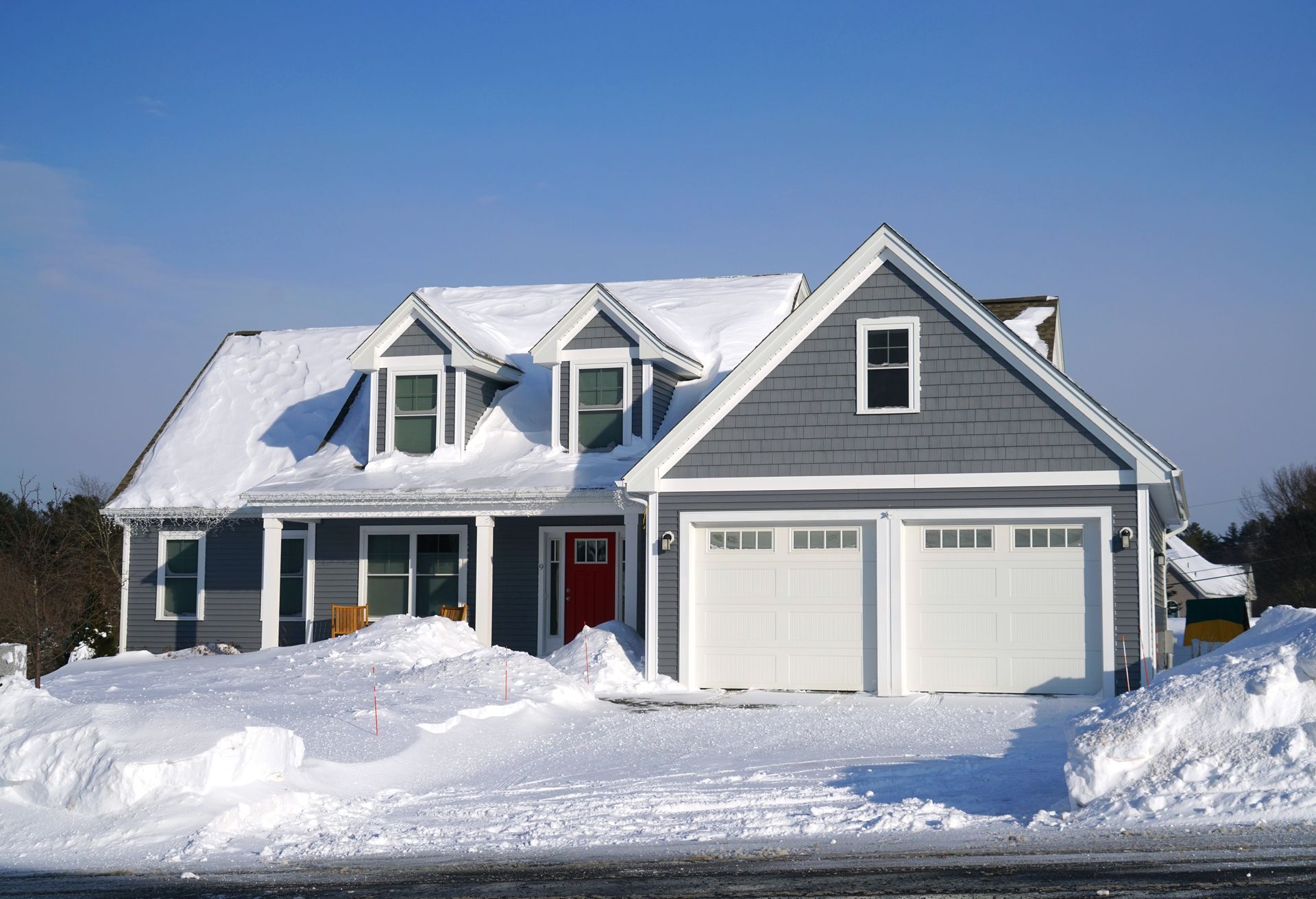 Gray house covered in snow with white garage doors and a red front door, under a blue sky.