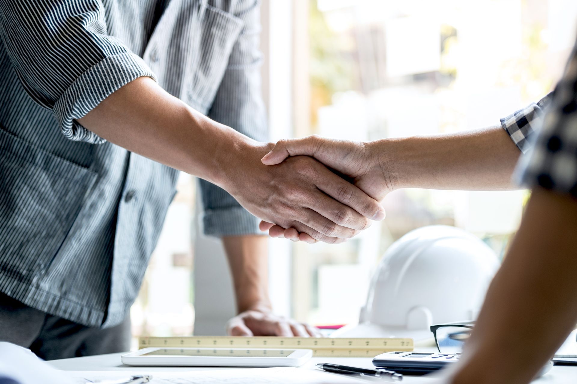 Two people shaking hands over a desk with blueprints and a hard hat.