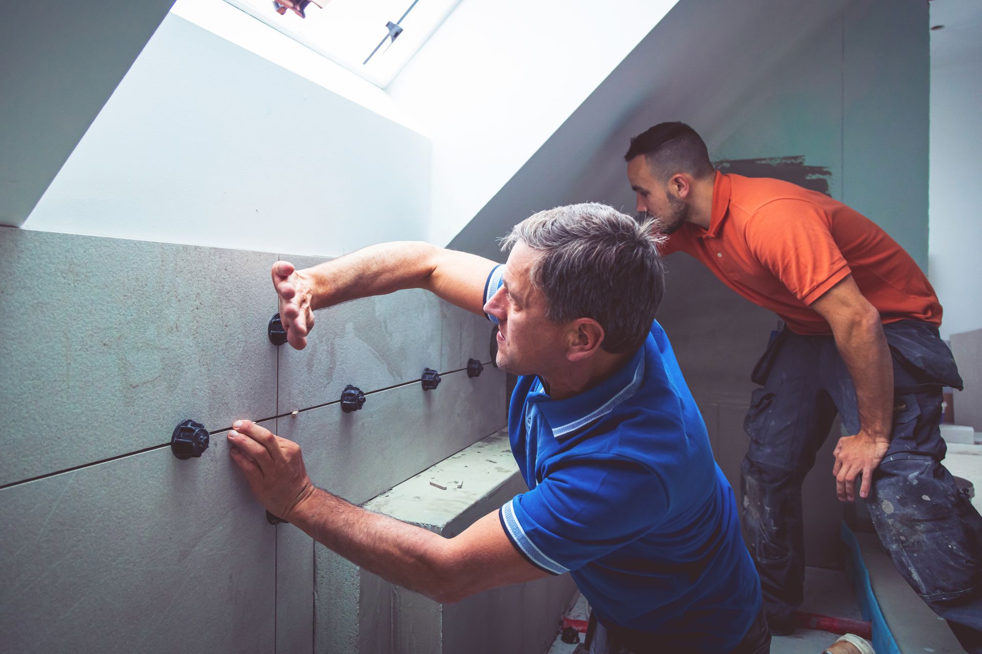 Two people installing wall panels in a room with a skylight; one man tightens fasteners.