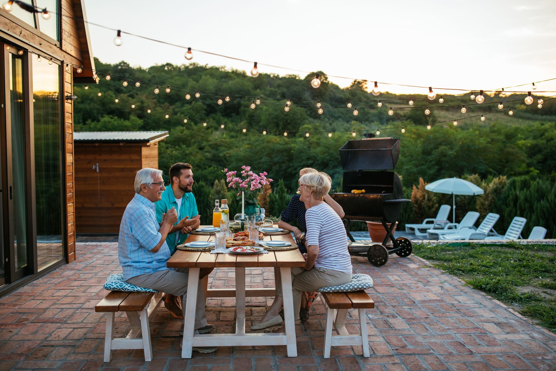 Family dining outdoors at sunset, seated around a wooden table. Grill and string lights in the background.