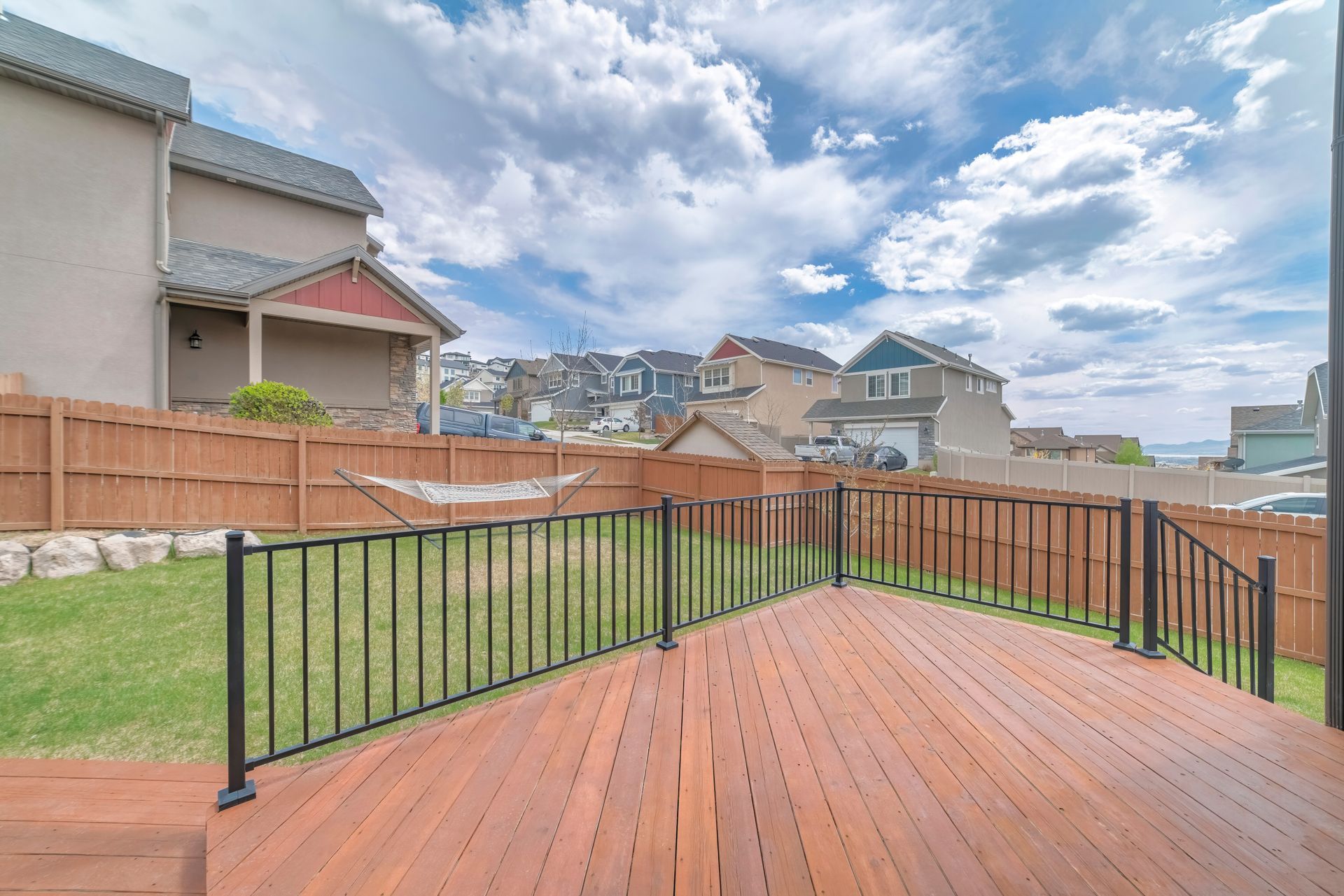 Wooden deck with black railing, grass, and wooden fence. Houses and blue sky in background.