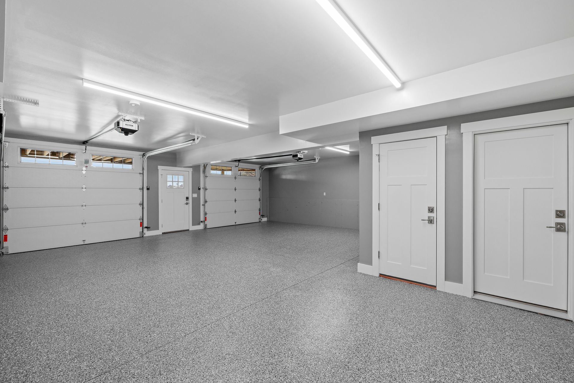Empty, brightly lit garage with gray speckled floor, white doors, and garage doors.