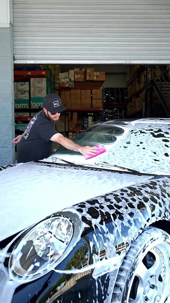 Auto detailer hand-washing the windshield and roof of a foam-covered Porsche 911 as part of a full exterior detail and paint correction prep.