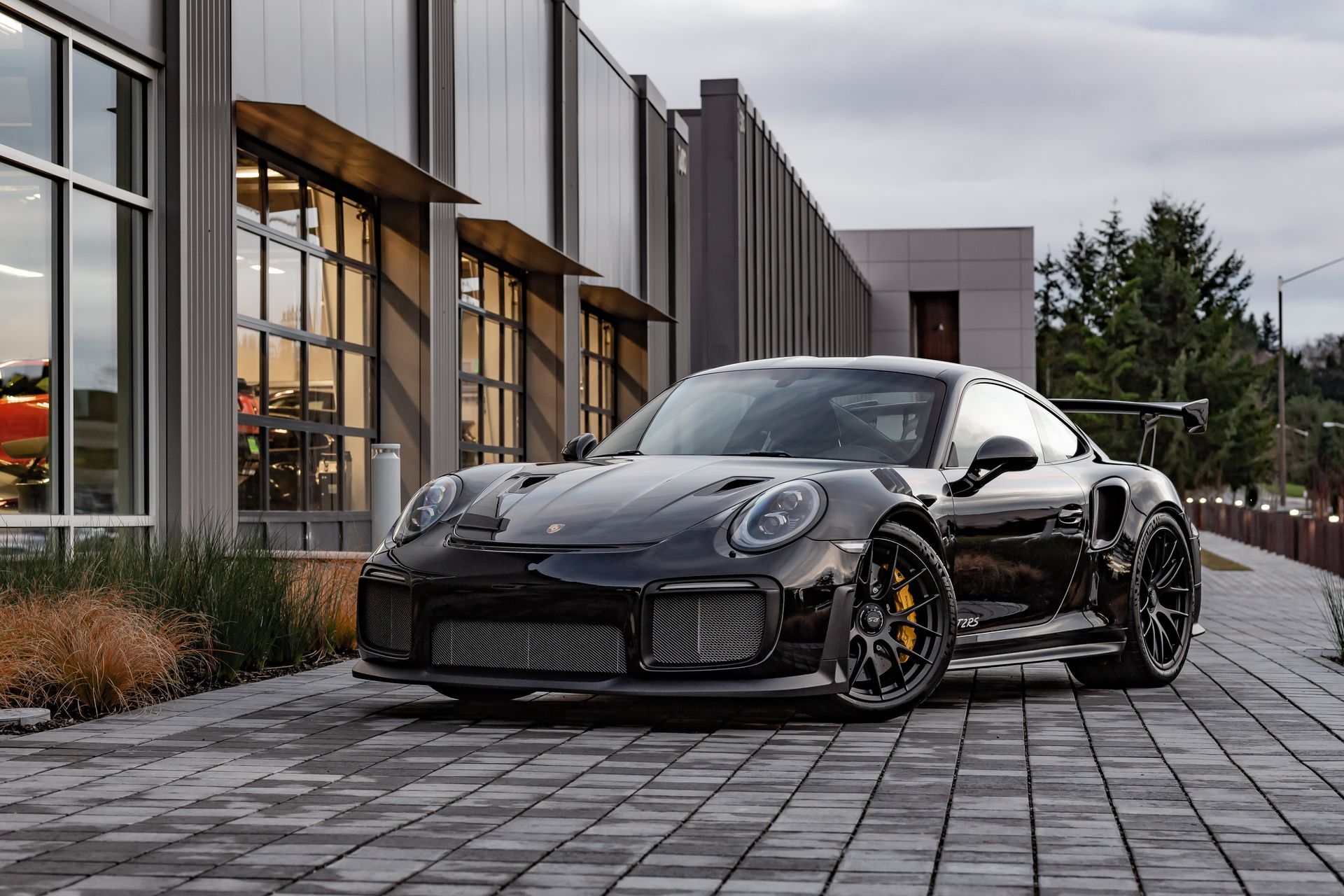 Black sports car parked on a paved driveway beside a modern glass building at dusk