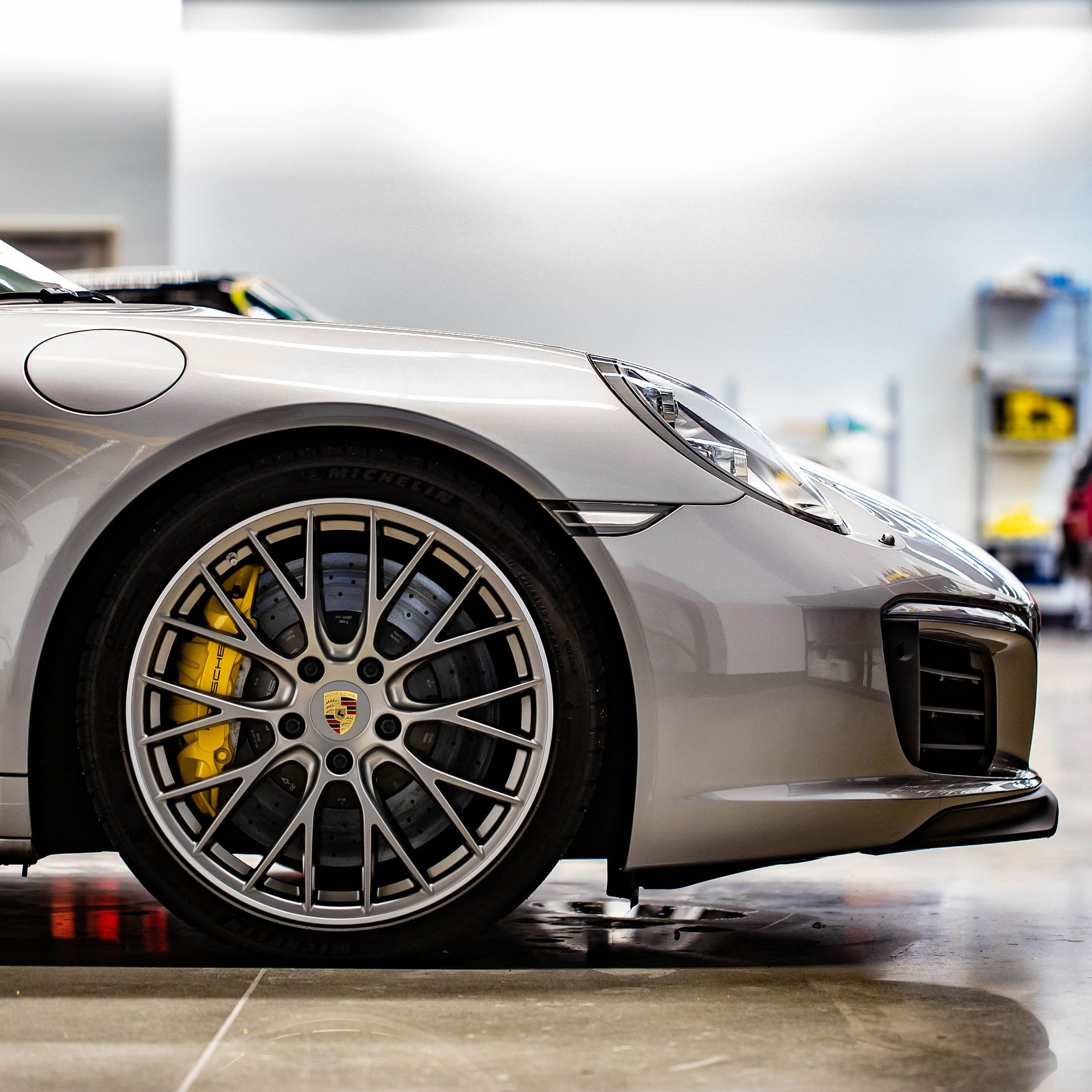 Silver Porsche sports car close-up, showing front wheel and bumper in a garage with blurred background