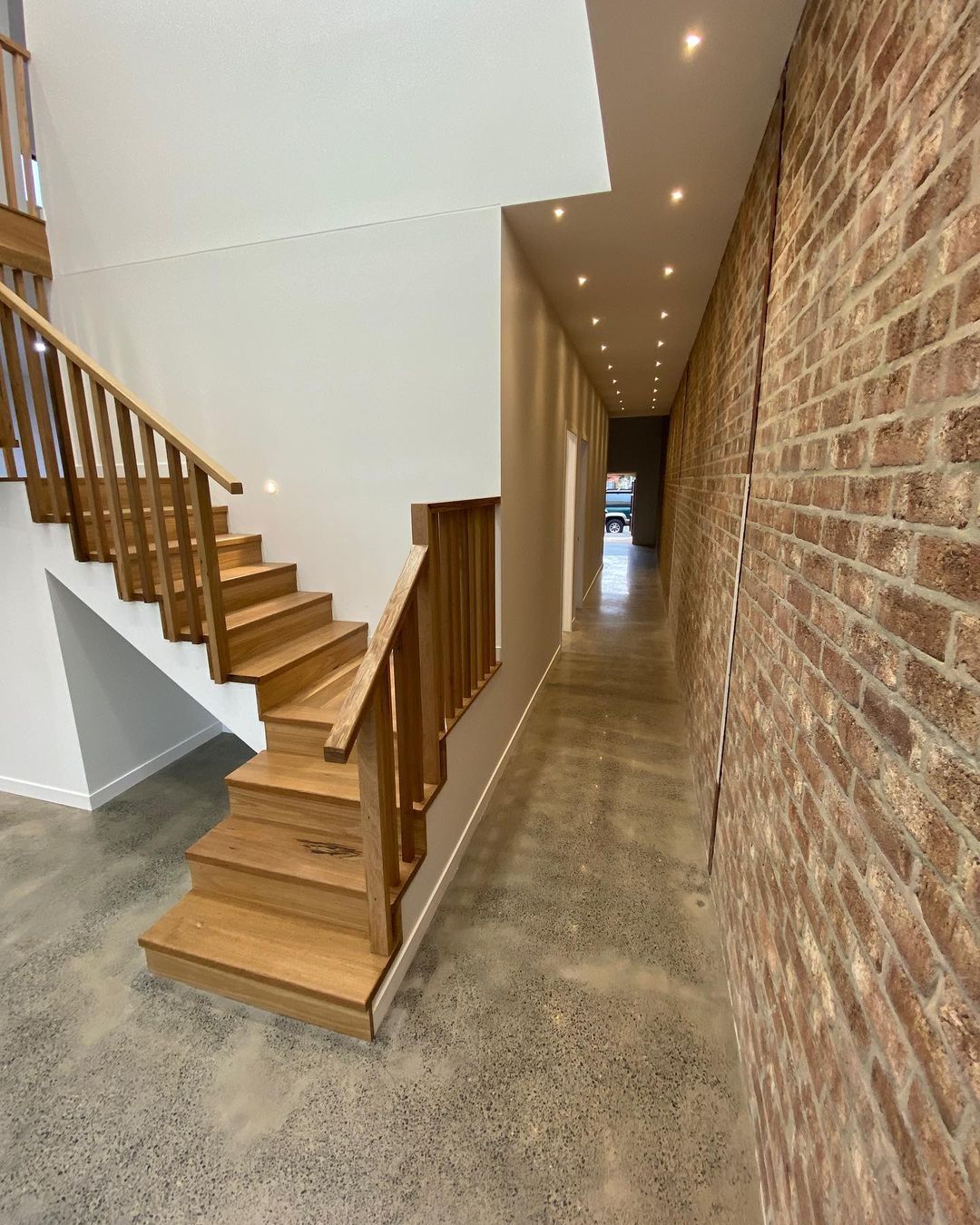 A long hallway with a wooden staircase and a brick wall.