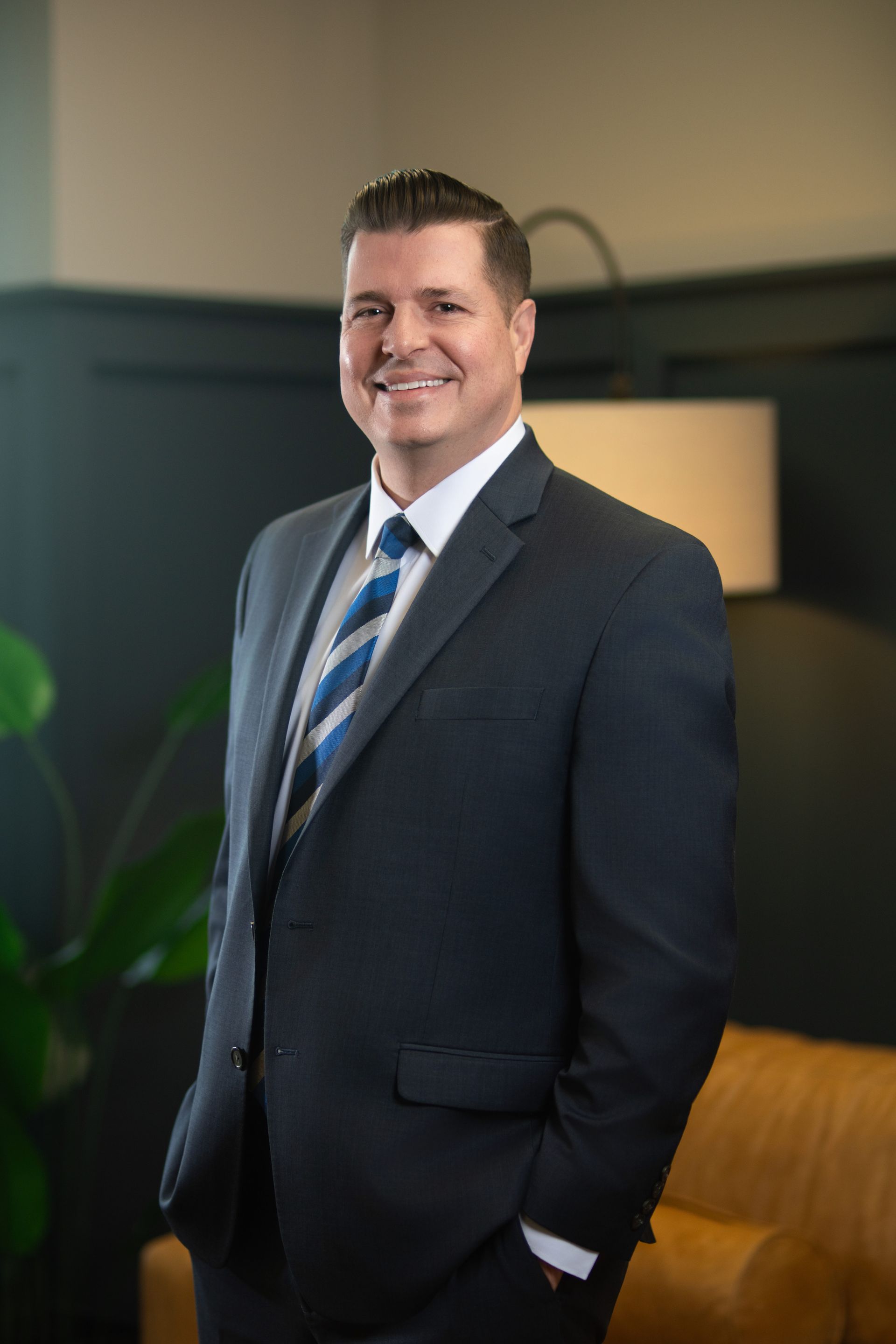 Man in a dark suit and blue striped tie standing in an office, smiling near a lamp and couch.