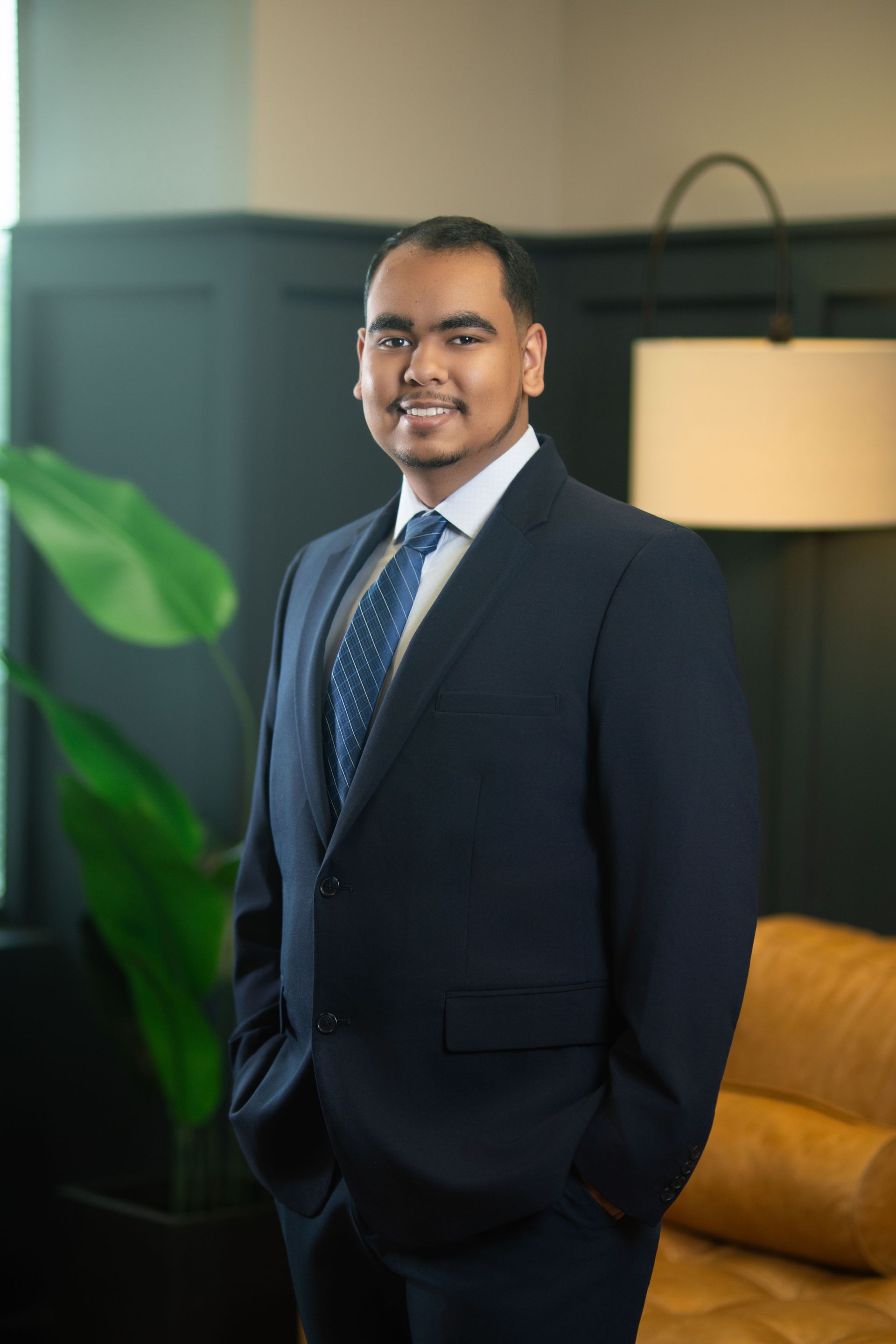 Man in a navy suit standing indoors beside a mustard sofa and floor lamp, smiling at the camera