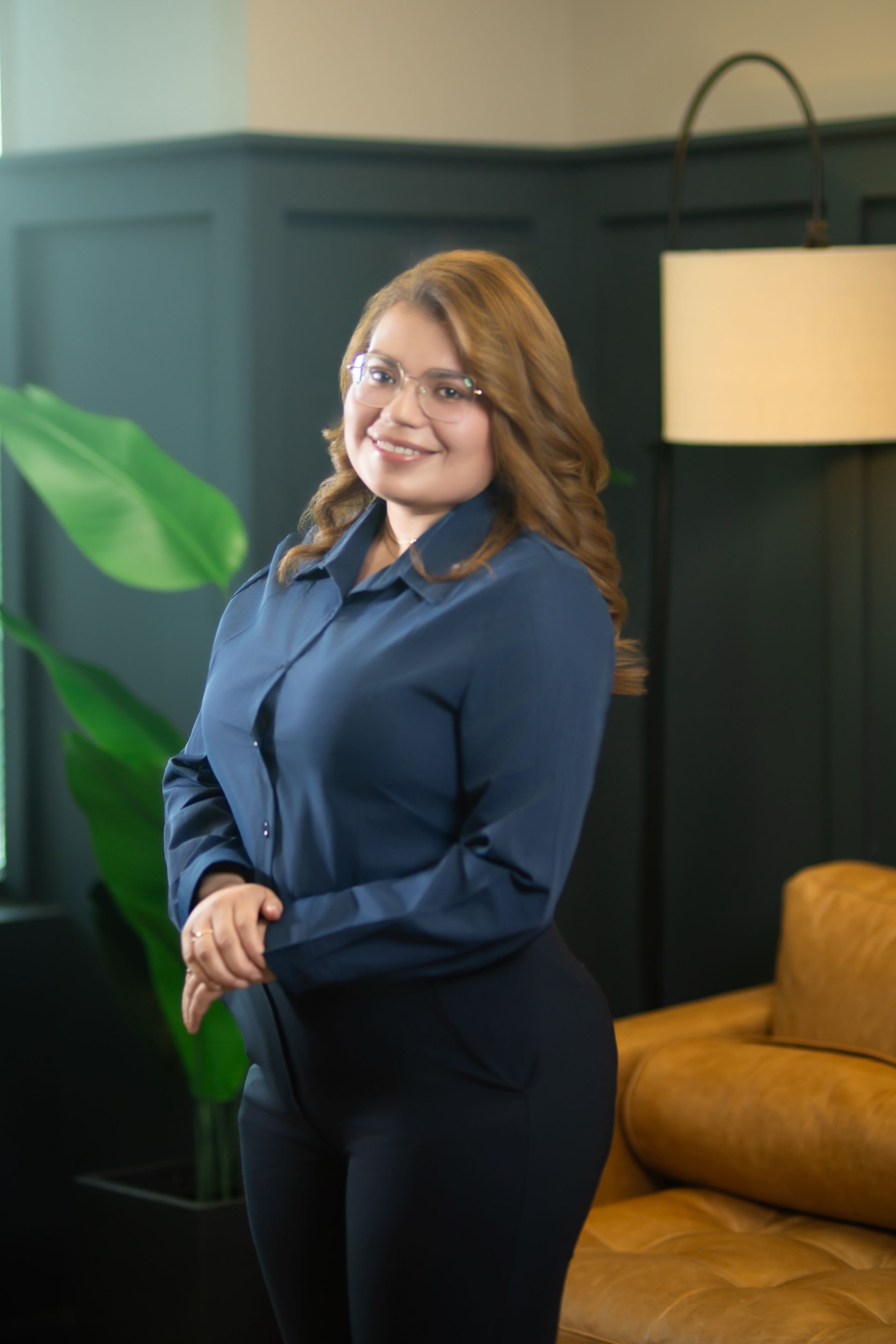 Woman in a navy blouse and black pants smiling in a modern room with a green plant and tan chair.