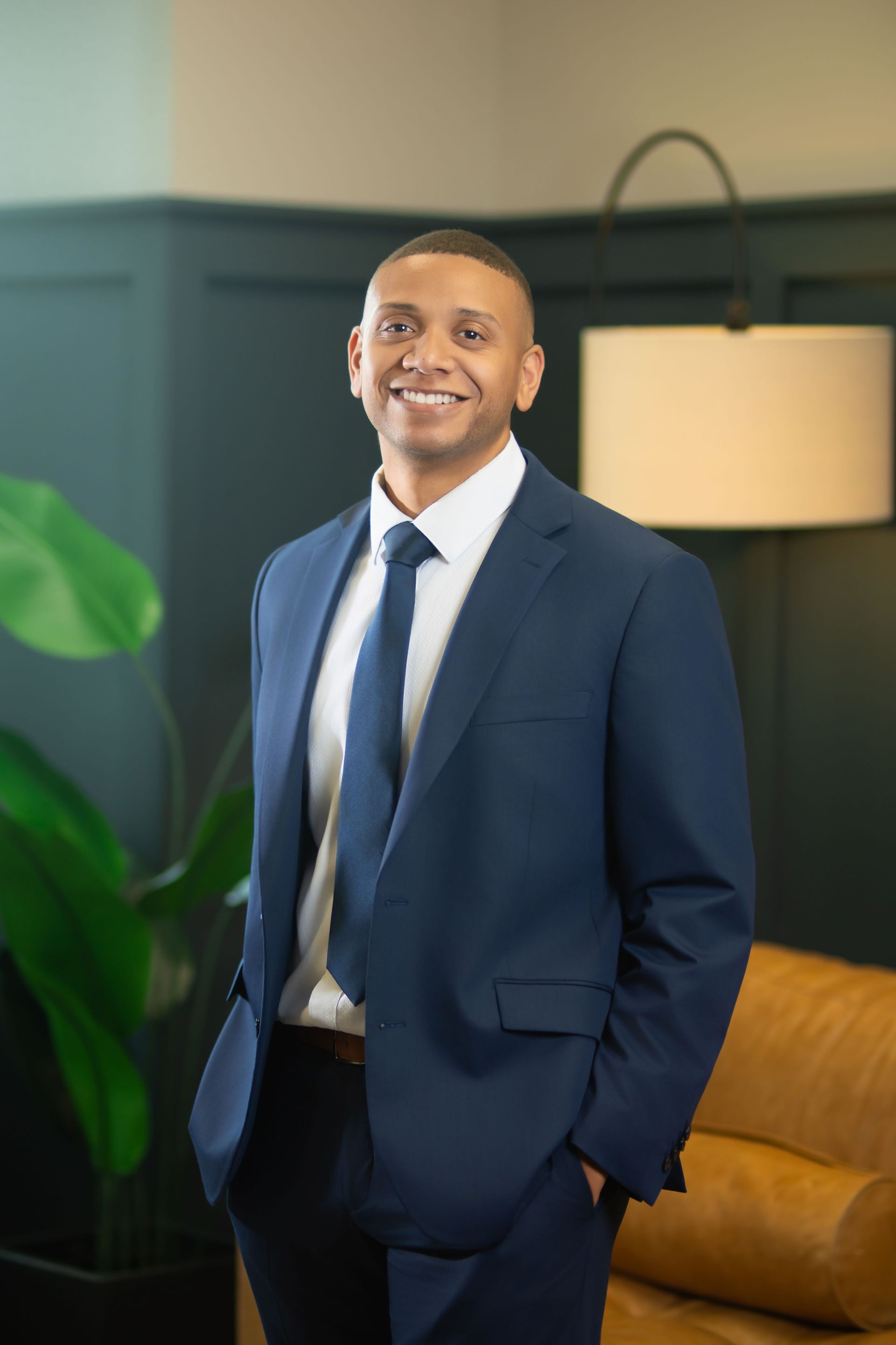 Smiling man in a navy suit standing in a modern office lounge with a lamp and mustard sofa.
