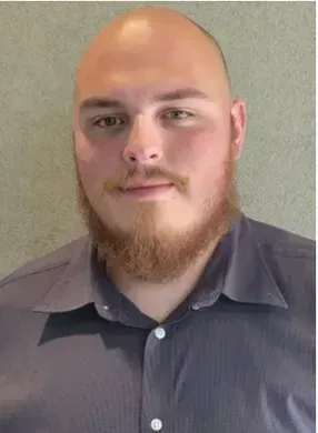 Portrait of a bald person with a reddish beard wearing a gray button-up shirt against a plain wall