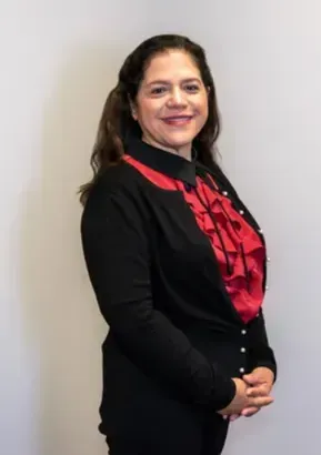 Woman in a black blazer with a red ruffled blouse, standing against a plain light wall and smiling.