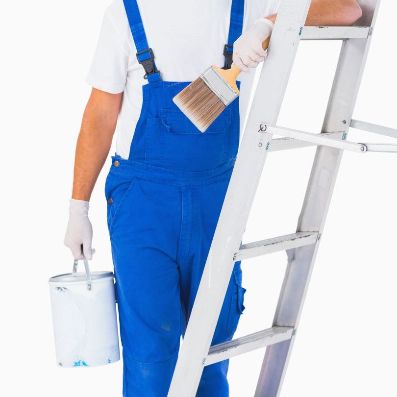 A man in blue overalls is standing on a ladder holding a paint bucket and brush