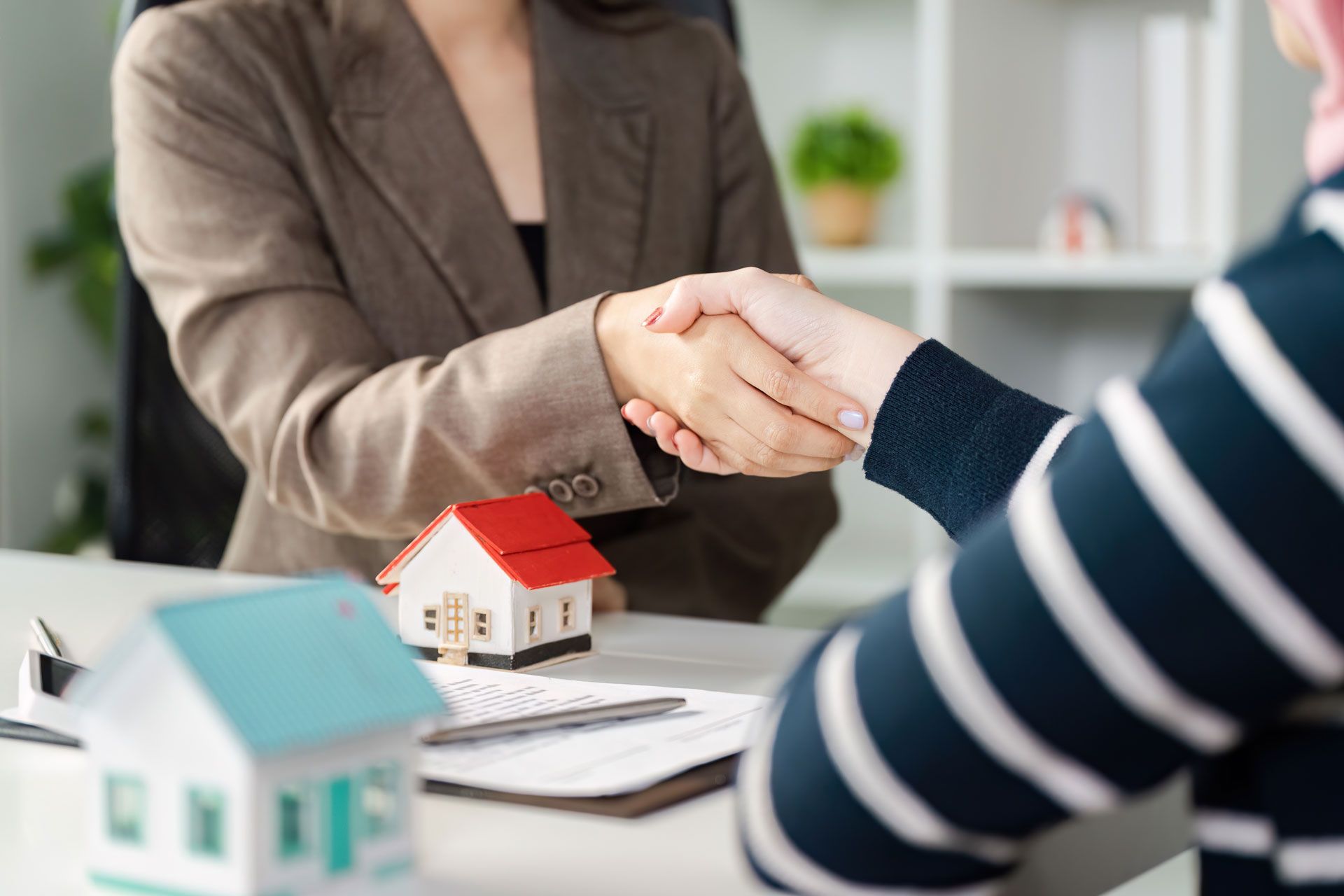 Two people shaking hands across a desk with miniature houses, suggesting a real estate agreement