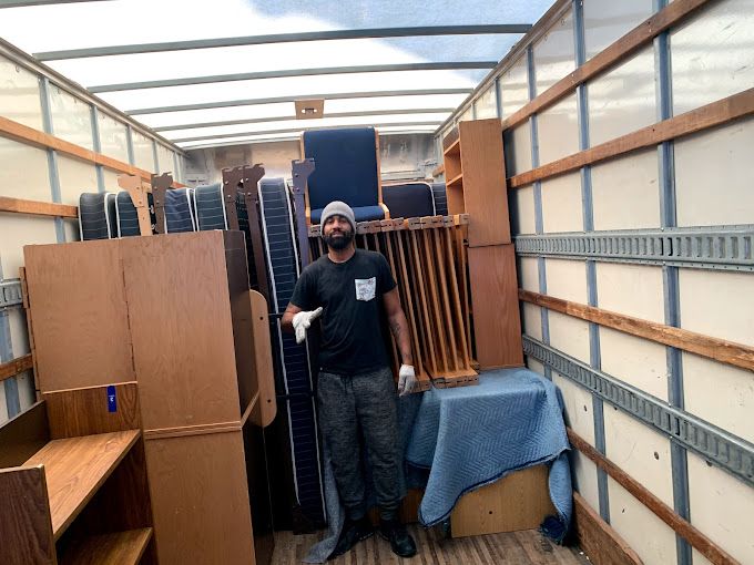 Man stands in a truck filled with furniture, including dressers, mattresses, and a table covered with a blue blanket.