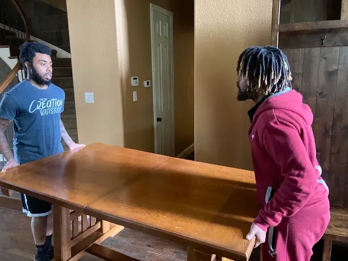 Two men lifting a large brown table indoors. One wears a gray shirt, the other a red hoodie.