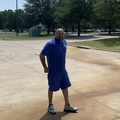 Man in blue outfit dances in a parking lot, trees in the background.