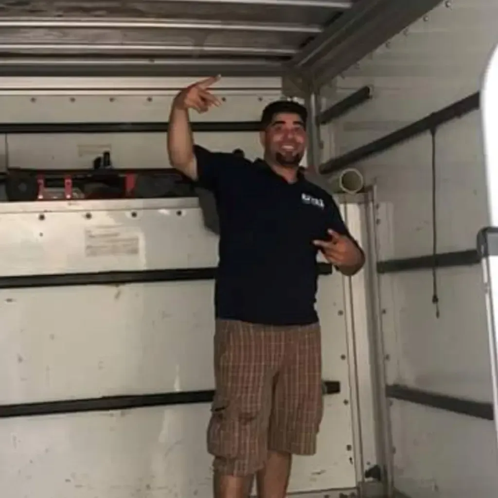 Man in moving truck, arms up, making peace signs, smiling. Wearing a blue shirt and brown shorts.