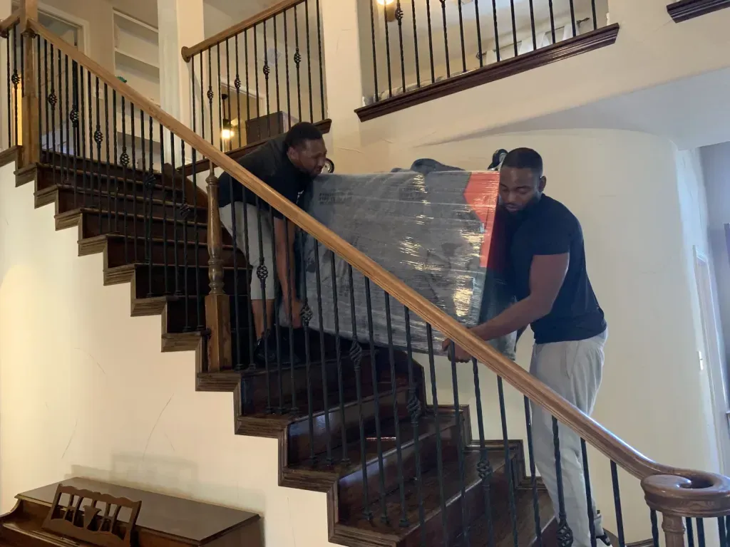 Two men carefully carry a wrapped object down a wooden staircase with a wrought iron railing.