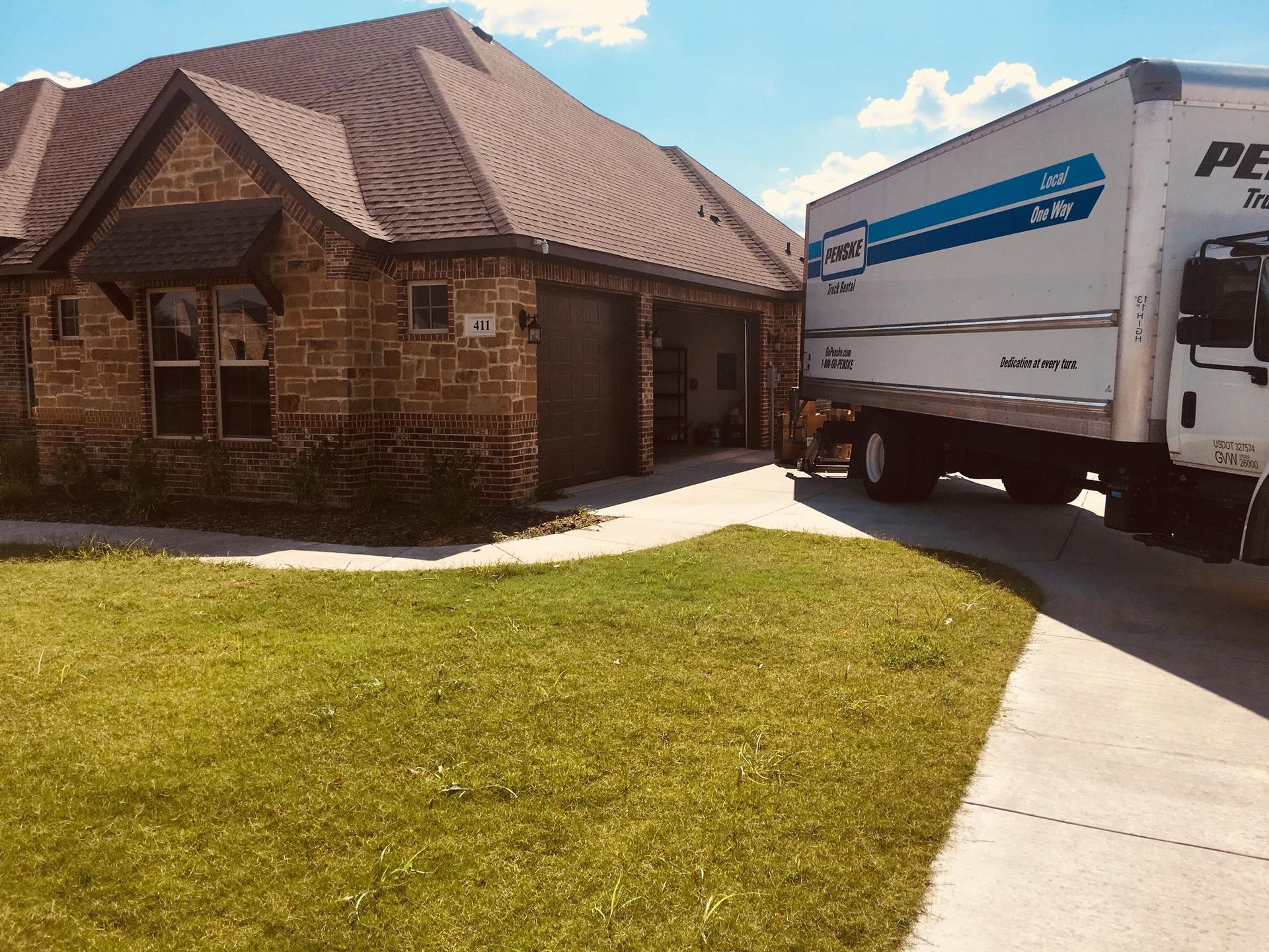 Moving truck parked in front of a brick house with a driveway and green lawn on a sunny day.