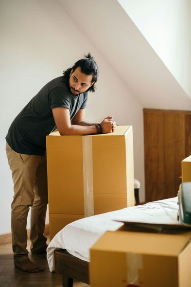 Man leaning on moving box in bedroom. He has dark hair and is wearing a grey shirt and tan pants.