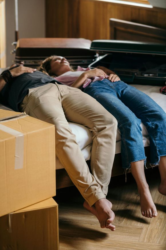 A couple rests on a bed surrounded by packed boxes and suitcases after a tiring move.