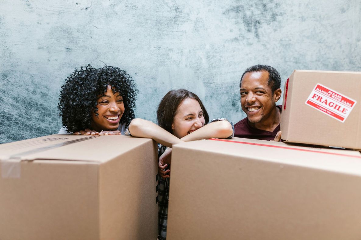 Three people smiling, leaning over cardboard boxes, moving or unpacking.