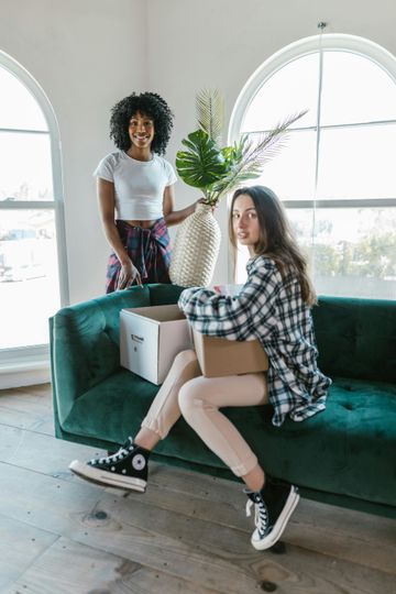 Two women unpacking in a new home. One on a teal couch with a box, the other by a vase, smiling.