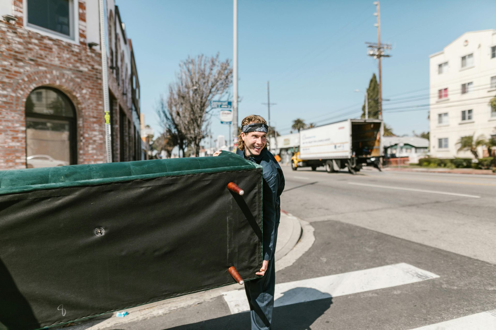 Person carrying a large, dark green couch across a street in front of buildings and a truck.
