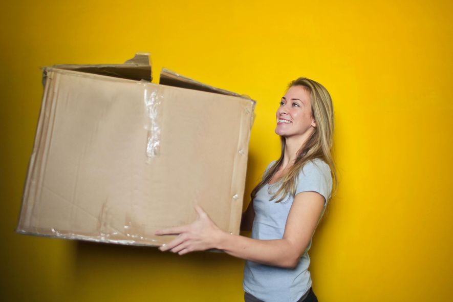 Woman with blonde hair smiling while carrying a large cardboard box against a yellow background.