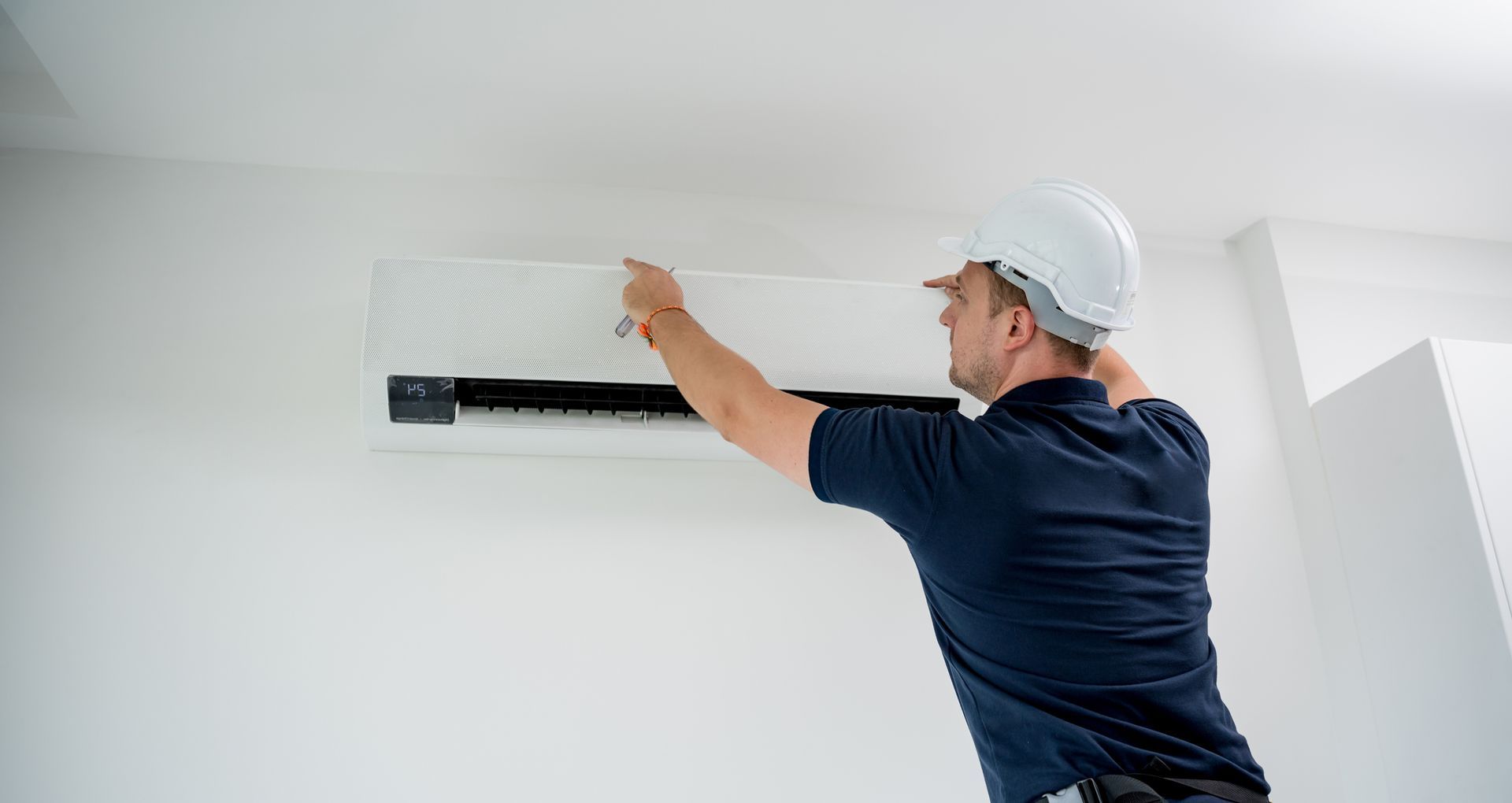 Worker in helmet adjusts wall-mounted AC unit in clean indoor space.