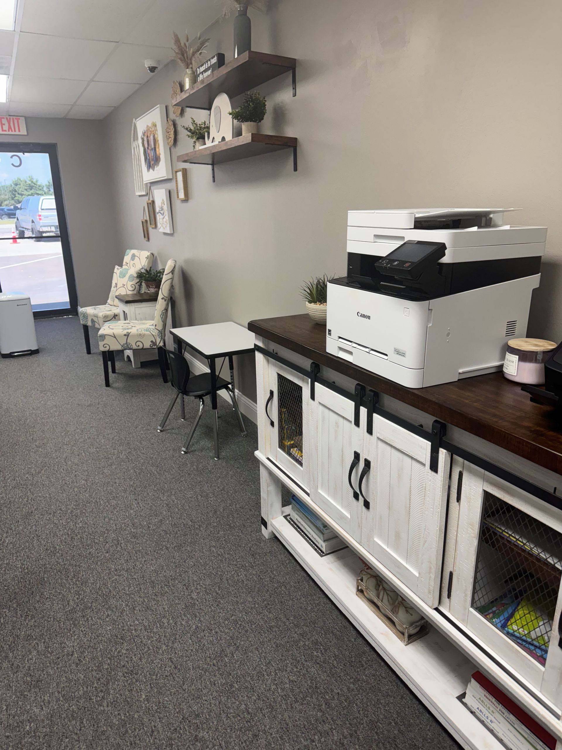 Office waiting area: white cabinet with printer, chairs, shelves with decor, gray carpet, and an open door.