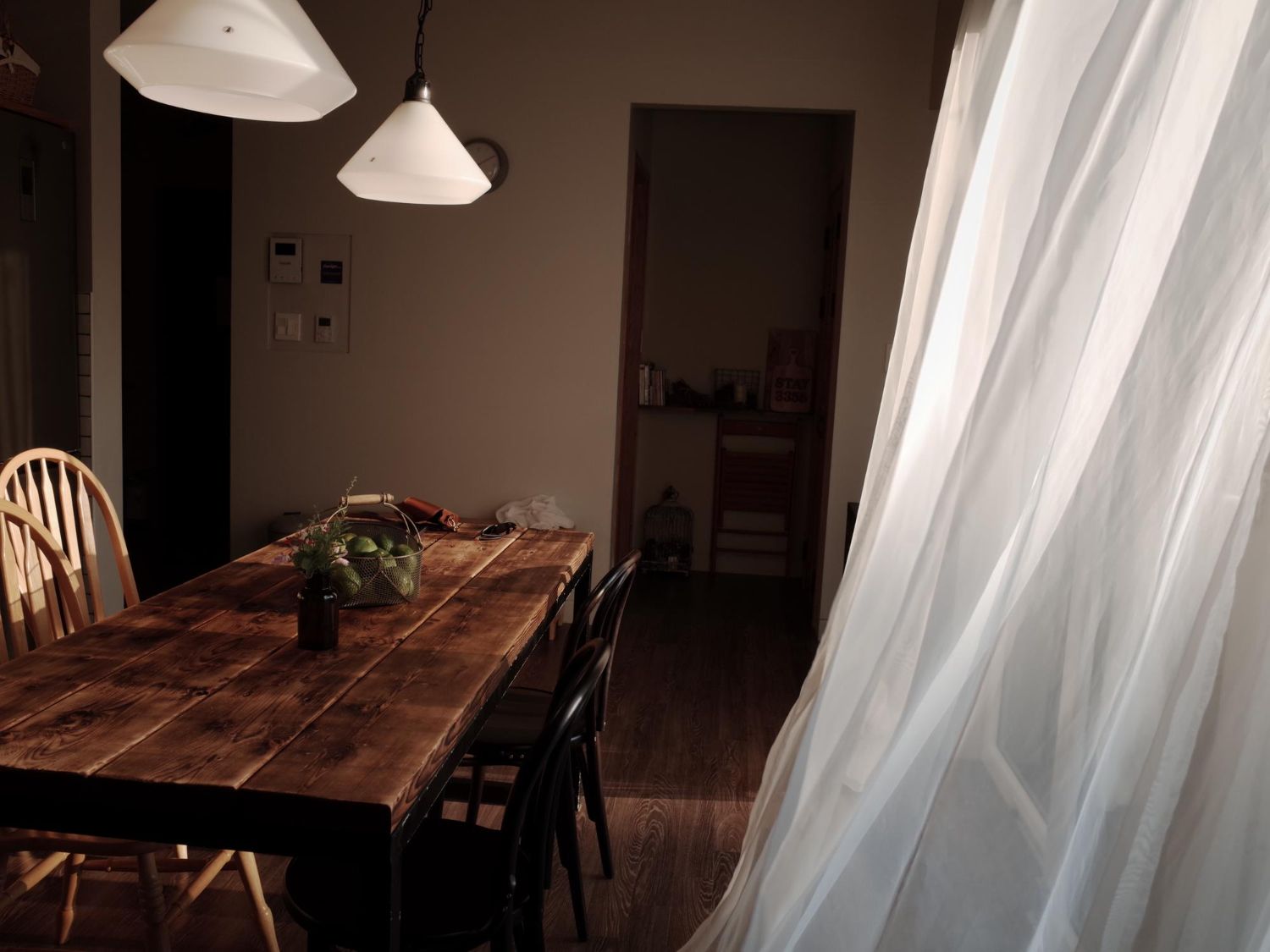 Dining room with wooden table, chairs, and sheer white curtains. Sunlight streams in.