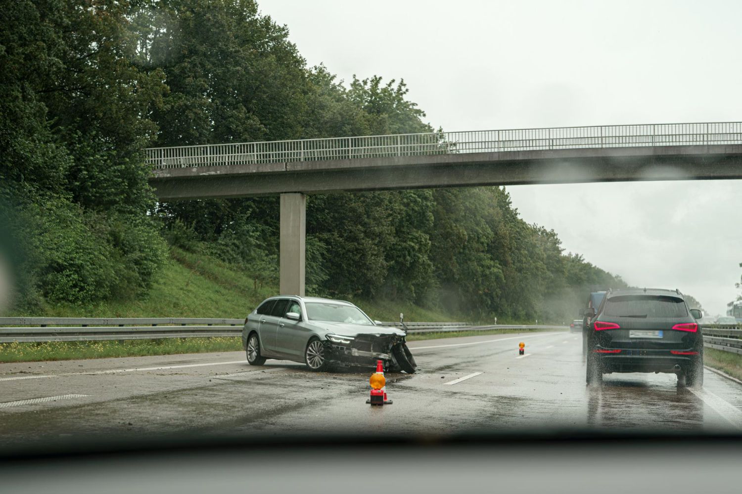 A silver car damaged in a traffic accident on a wet highway. A black SUV drives past.