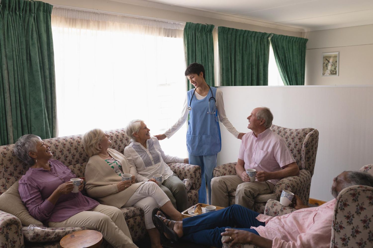 Nurse in scrubs chats with a group of seated seniors in a living room; green curtains.
