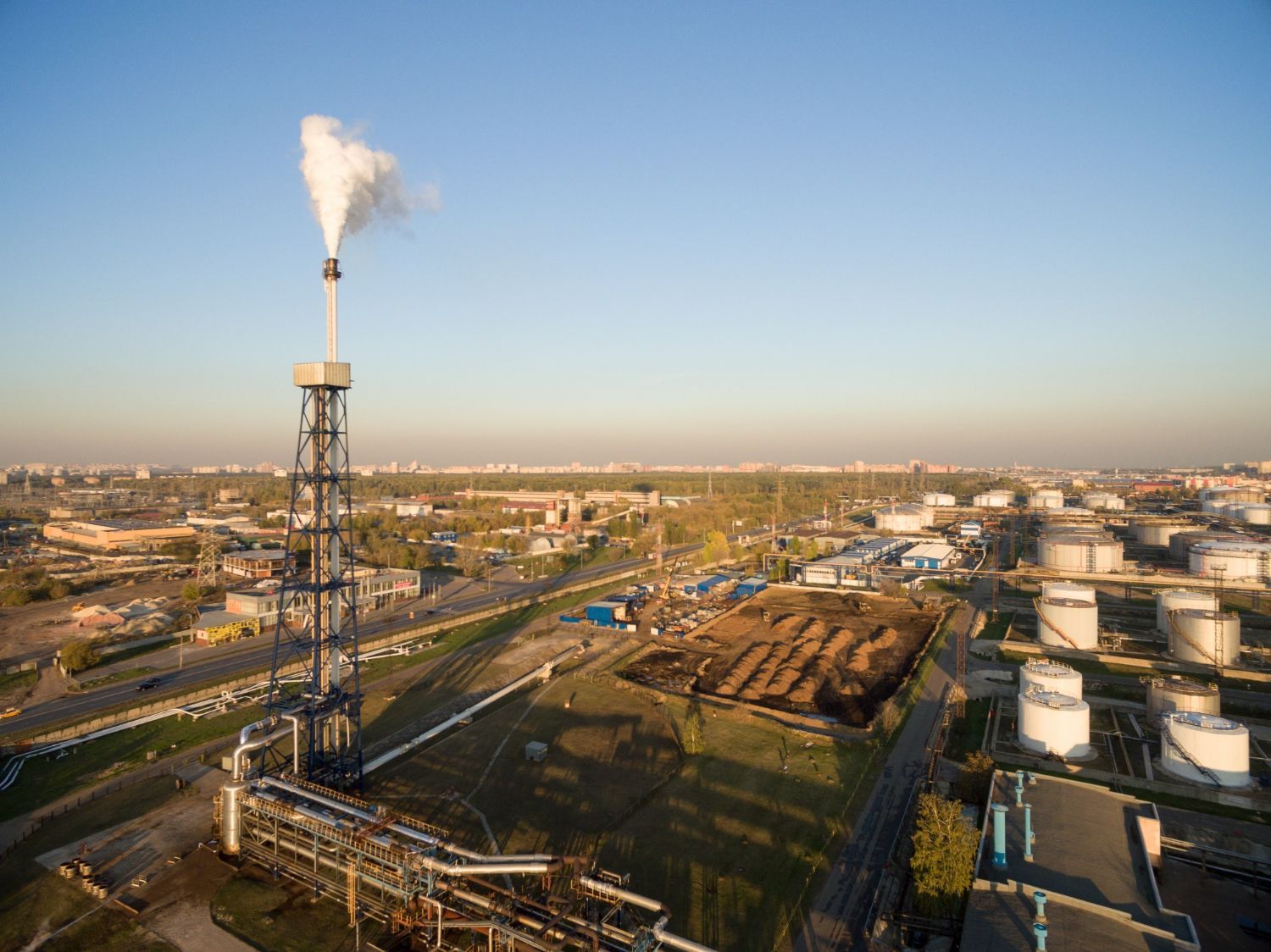 Industrial plant with smoke stack emitting steam, surrounded by storage tanks and buildings under a blue sky.