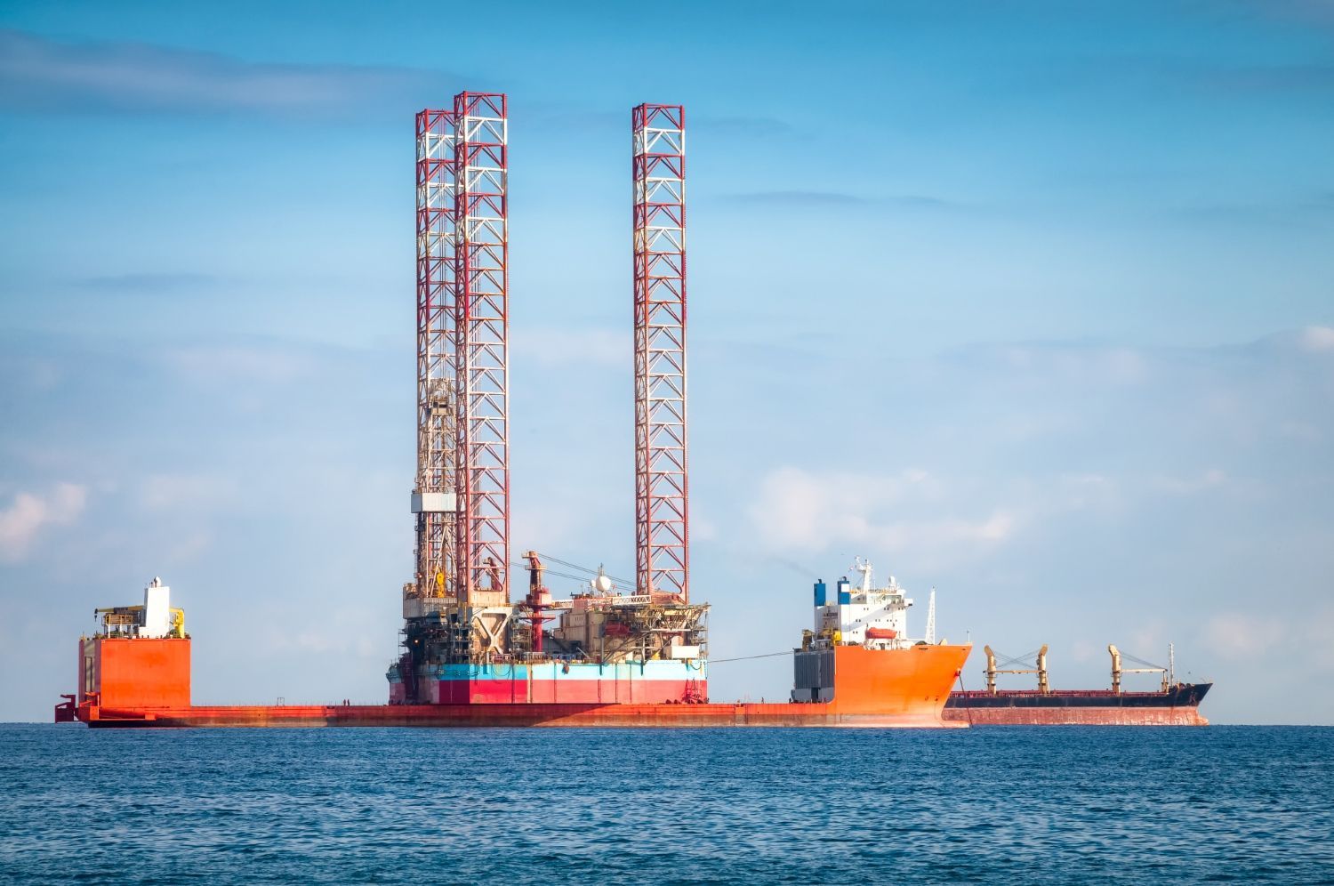 Offshore oil rig platform transported by a large orange ship, with two support vessels, on a calm blue sea.