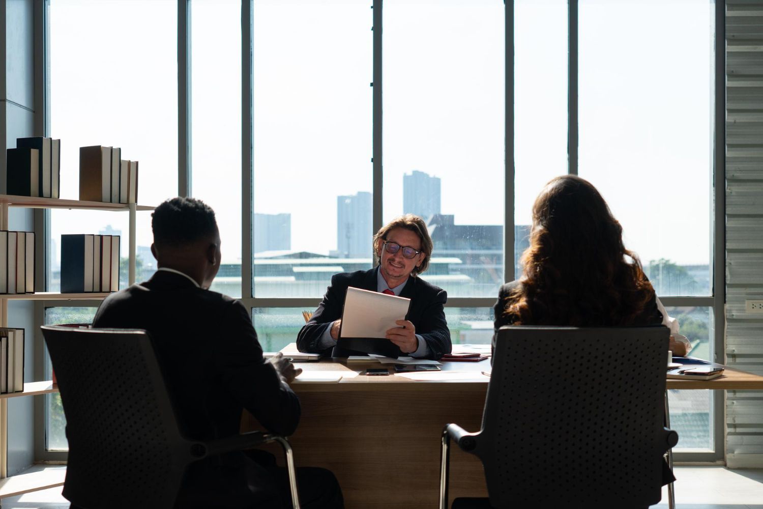 Three people at a desk in a well-lit office. One person is holding papers. Skyscrapers visible through a large window.