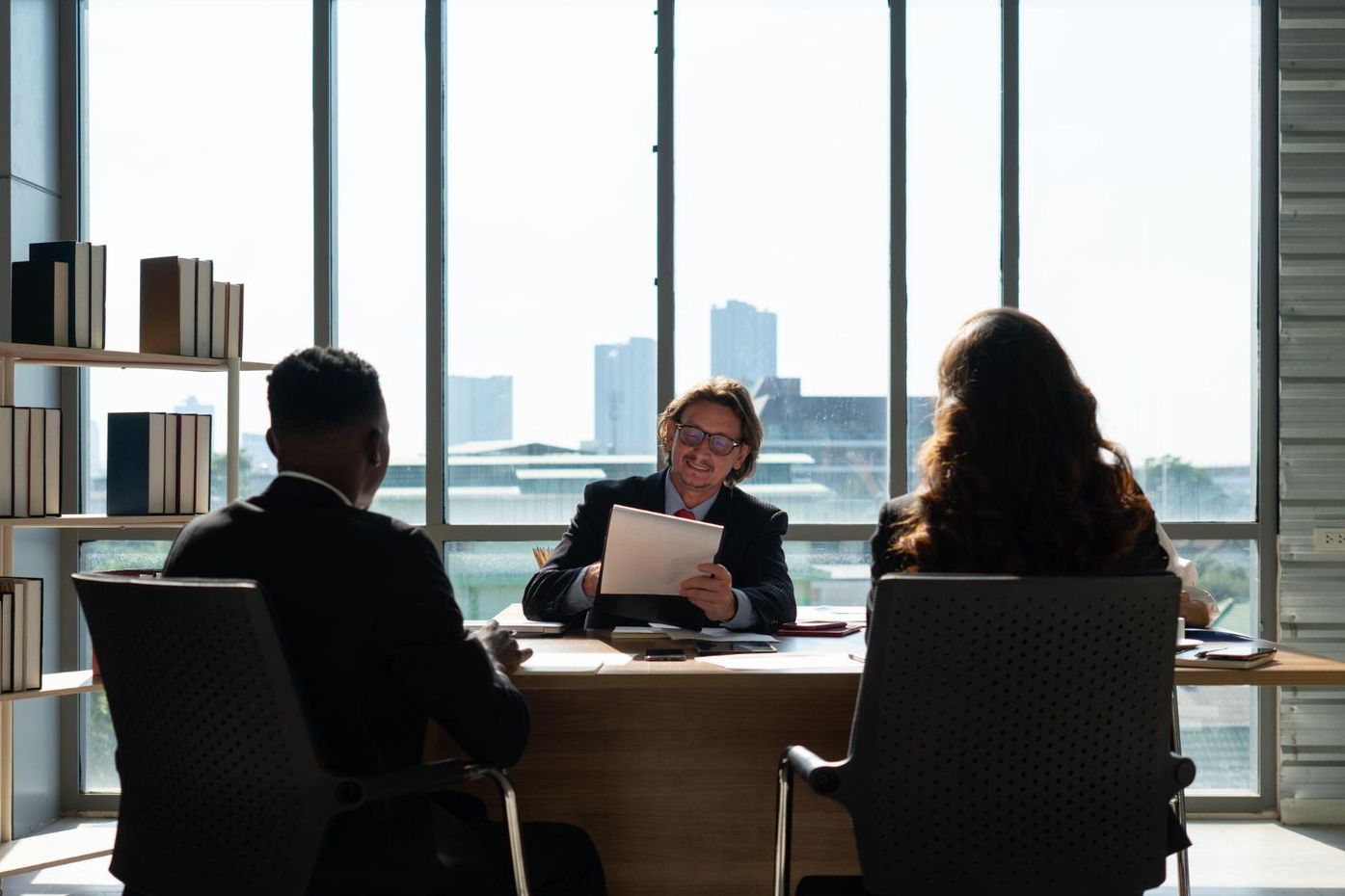 Three people at a desk in a well-lit office. One person is holding papers. Skyscrapers visible through a large window.