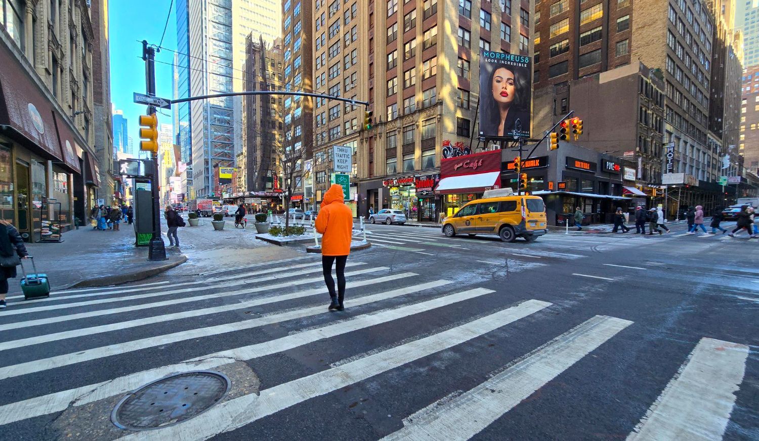 Person in orange jacket crossing a New York City street with crosswalks, buildings, and a yellow taxi.