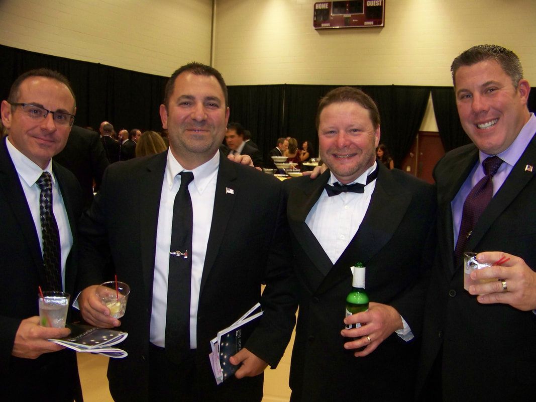 Four men in suits pose indoors, holding drinks, smiling. One in a tuxedo holds a bottle.