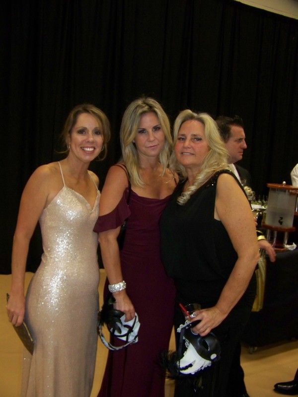 Three women in formal dresses pose, holding helmets. One in a sequined gown, one in burgundy, one in black. Dark background.